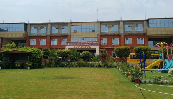Facade of a school building with a sign that reads Ayodhya, surrounded by a green lawn and shrubs; a colorful playground is visible on the right.