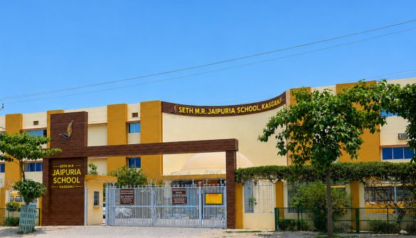 Seth M R Jaipuria School Kasganj - Jaipuria Schools - Jaipuria Schools Front view of a yellow-and-cream school building with a gated entrance and trees on a sunny day (Seth M.R. Jaipuria School).