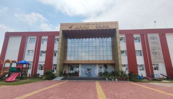 Front view of Jaipur School building with red side wings, a glass central entrance, and a playground visible on the left.