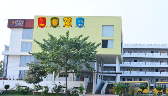 Seth M R Jaipuria School Lakhimpur - Jaipuria Schools - Jaipuria Schools School building with four crest emblems on a yellow wall, a tree in front, and a yellow bus parked to the right.