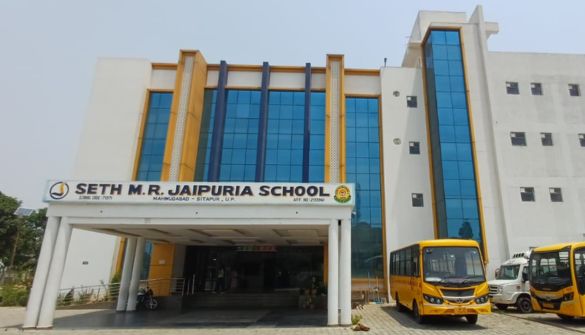 Front view of Seth M.R. Jaipuria School building with glass blue windows and a yellow school bus parked at the entrance