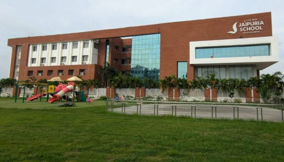 Seth M R Jaipuria School Shahjahanpur - Jaipuria Schools - Jaipuria Schools Front view of Jaipuria School campus: red-brick building with a glass atrium, and a playground on the grassy foreground.