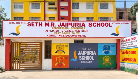 Front view of Seth M.R. Jaipuria School building with yellow and red facade, large sign, and colorful entrance banners crisscrossing the facade.