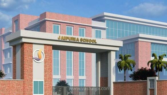 Front view of Jaipuria School building with brick exterior, tall glass windows, a porte-cochère entrance, the school name on the facade, and palm trees against a blue sky.