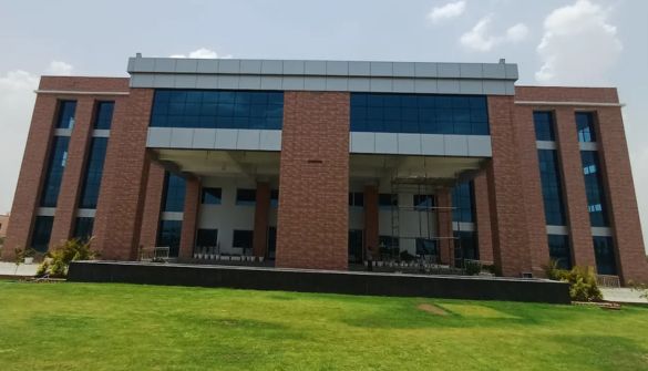 Brick campus building with a large covered entrance and blue-tinted windows, set on a well-kept lawn on a sunny day.